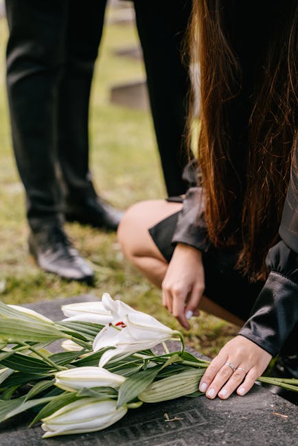 A woman dressed in black, kneeling on the ground at a graveside, arranging a bouquet of white lilies with long green leaves, possibly preparing a floral tribute. Her long brown hair partially obscures