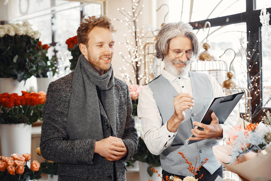 A male customer with curly brown hair and a light beard, dressed in a dark grey coat and scarf, stands inside a flower shop with bright natural light from large windows. Beside him, a floral specialis