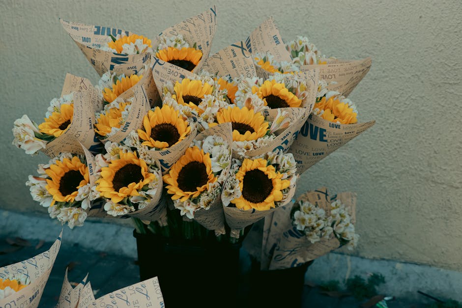 A bouquet of fresh sunflowers with bright yellow petals and dark centers, arranged with smaller white and light yellow flowers, wrapped in patterned paper with words like 'love' and 'joy' printed on i