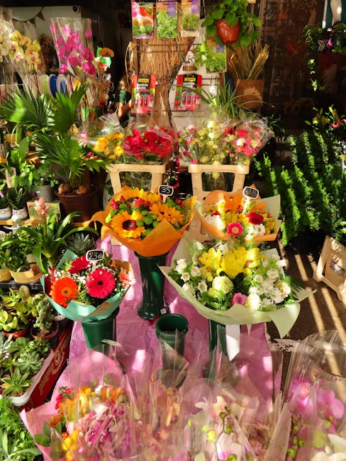 A vibrant display of fresh flower bouquets arranged on a pink table in a plant shop, featuring a variety of colorful blooms including bright red, yellow, pink, and white flowers. The bouquets are wrap
