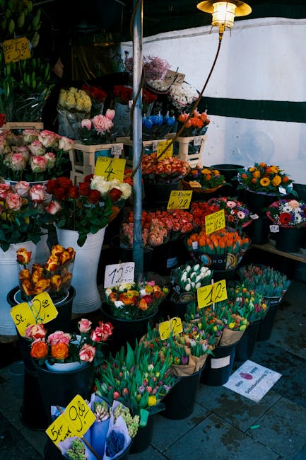 An outdoor display of fresh flowers at a market stall, featuring a variety of vibrant blooms including pink roses, orange tulips, yellow daisies, and multicolored carnations arranged in black pots and